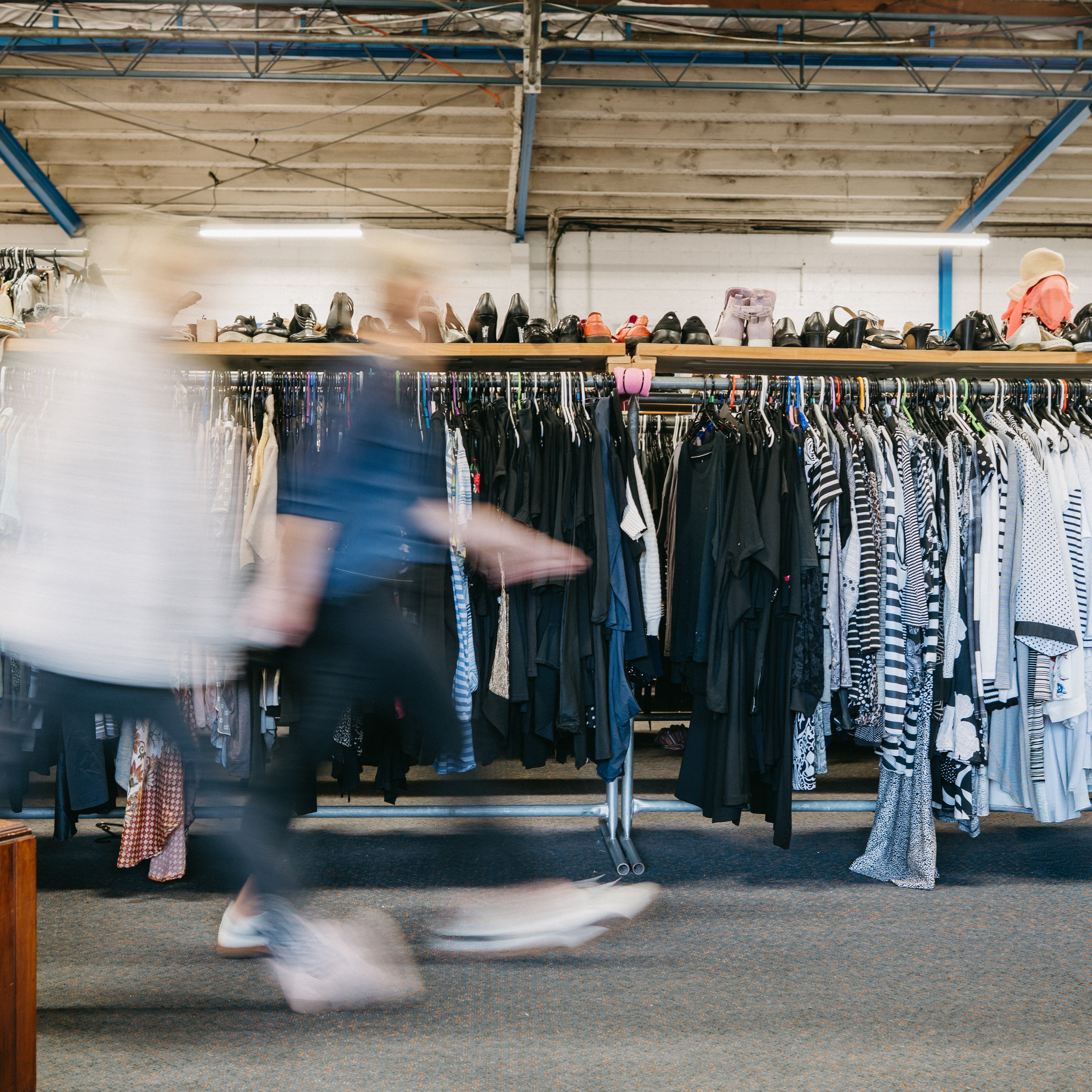 Blurred shoppers move past rows of clothing racks in a secondhand or thrift store.