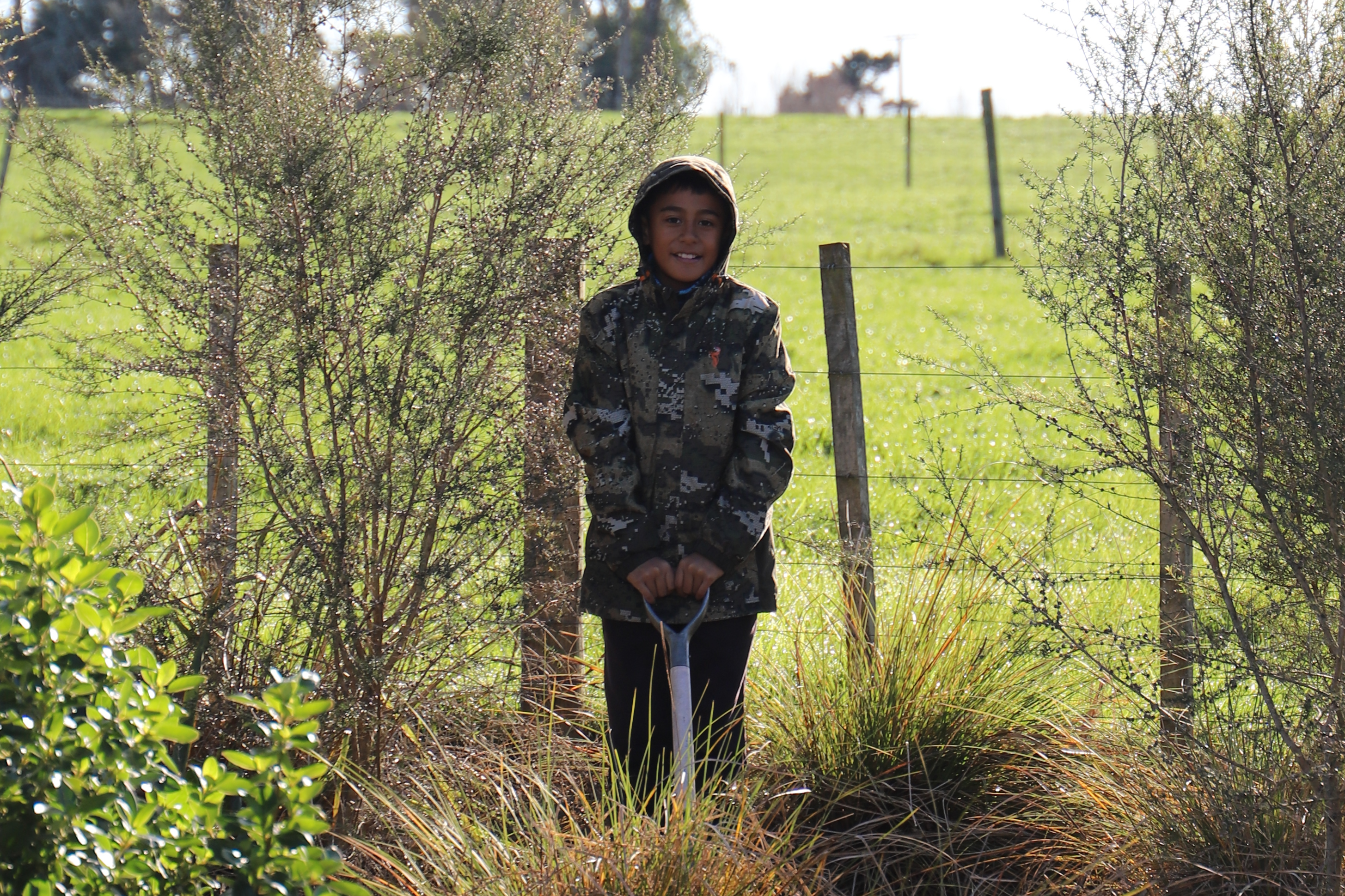 A child in a camo jacket stands in front of a wire fence, holding a shovel. The background shows green fields and shrubbery under a sunny sky.