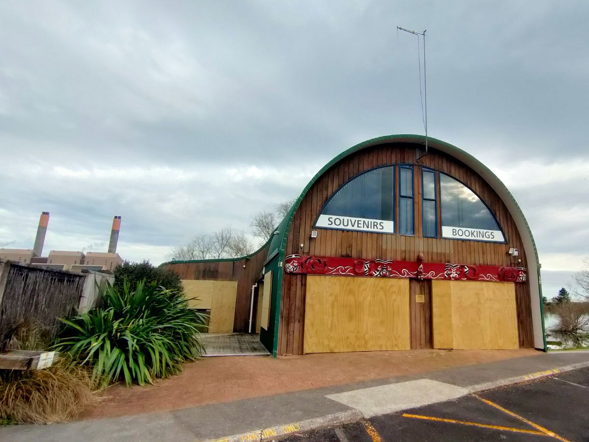 Curved wooden building with "Souvenirs" and "Bookings" signs, adorned with red carvings. Surrounded by a cloudy sky and lush greenery.