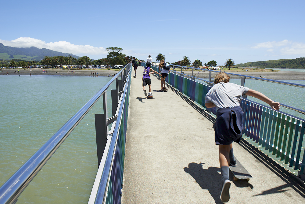 Kids joyfully skateboarding on a sunny bridge over turquoise water, with a background of palm trees, beachgoers, and distant green hills.