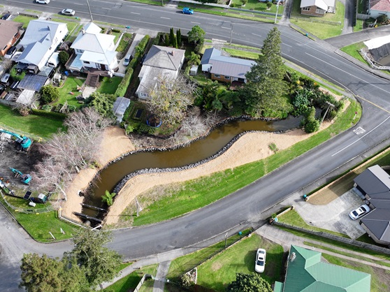 Aerial view of a landscaped suburban area featuring winding creek lined with rocks, surrounded by neatly arranged houses and lush greenery. Calm atmosphere.
