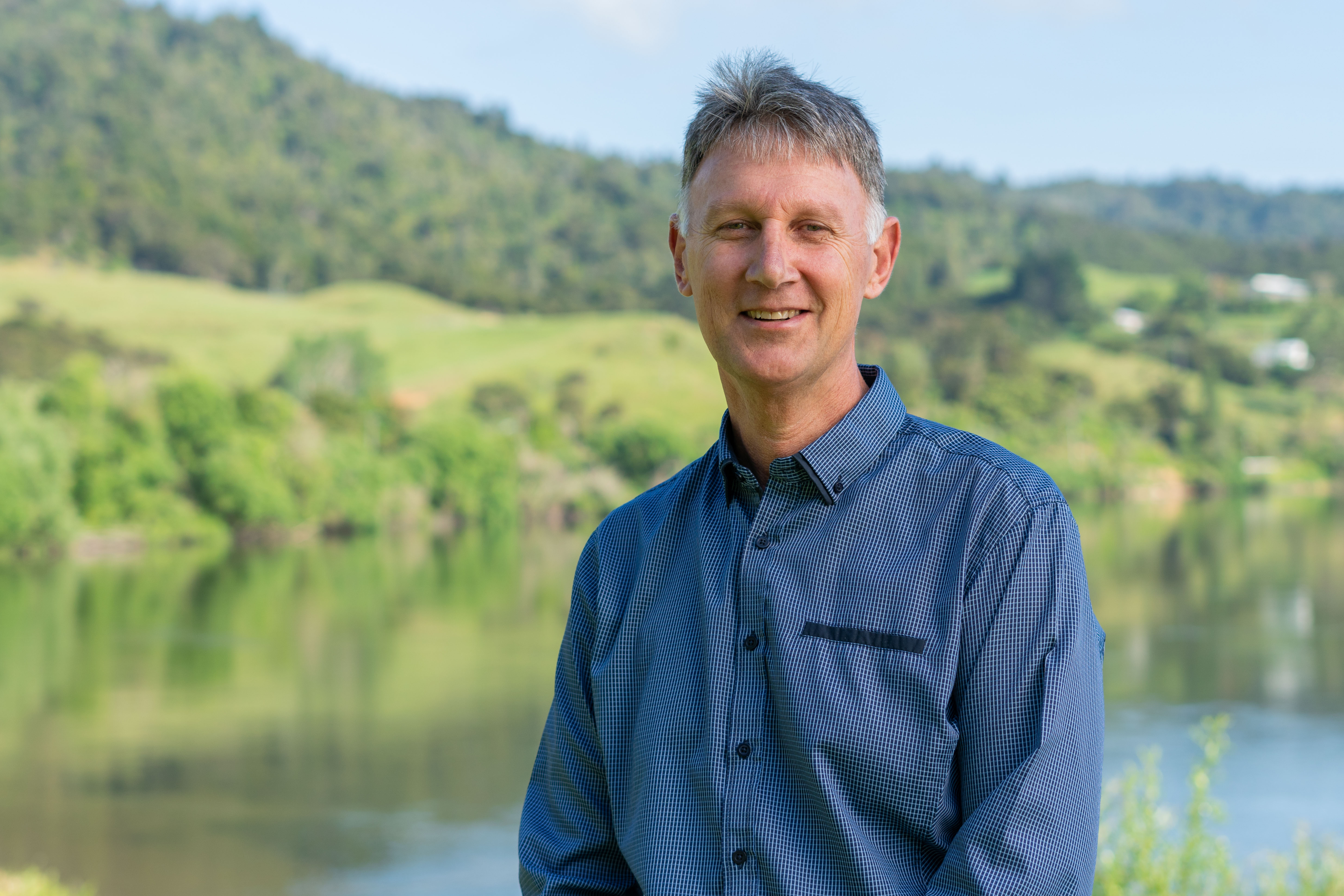 A man stands smiling in a blue checkered shirt against a lush green landscape with a river and hills under a clear sky, conveying a serene tone.